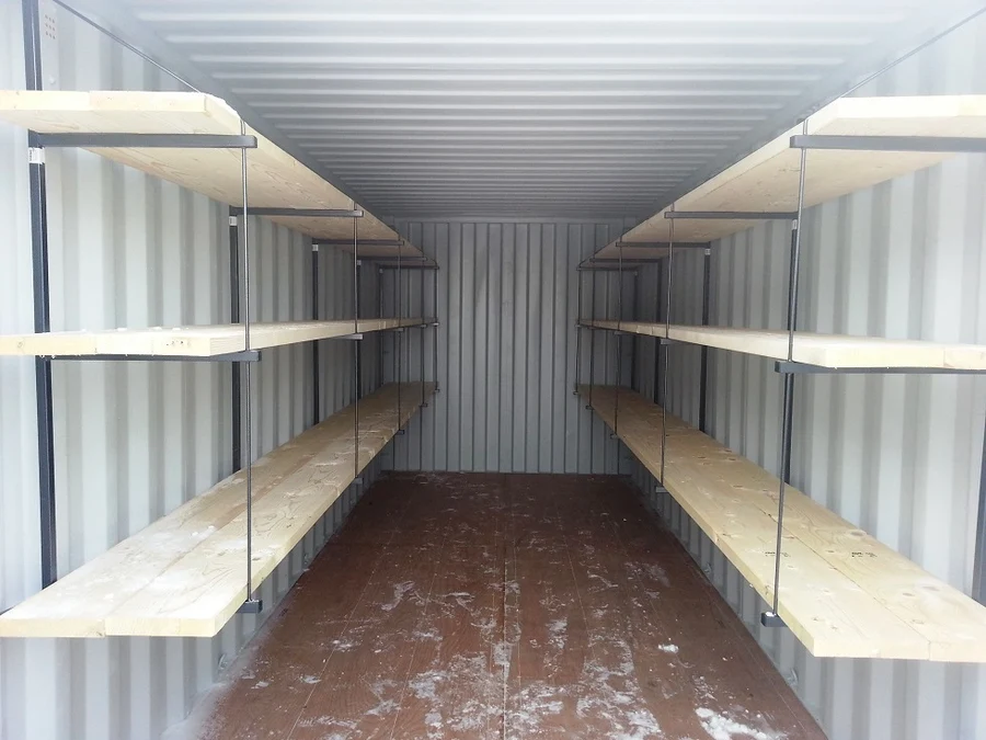Three-tier shelving brackets with lumber shelves installed along both walls of a 20ft shipping container