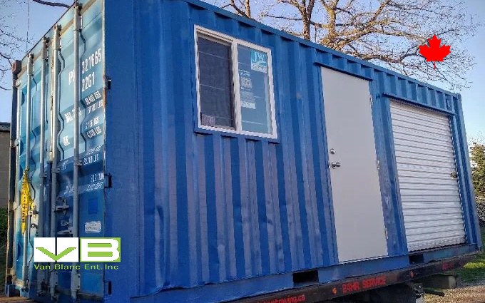 A blue converted unit with a window, man door, and roll-up riding a trailer up a lane under bare trees.