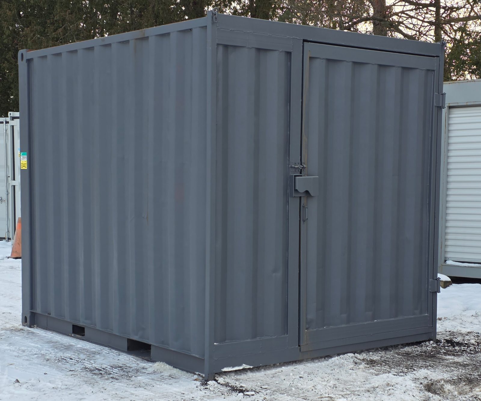 Repainted grey 10 foot container with a wide single man door and a padlock hasp, fork pockets showing along the base, standing on packed snow