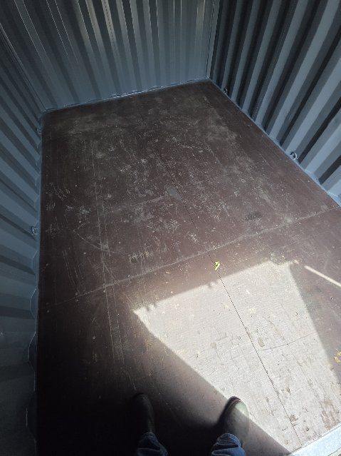 Looking straight down at the scuffed wood floor of the 12ft, with seams between the sheets, sunlight across one corner and a pair of boots at the bottom edge