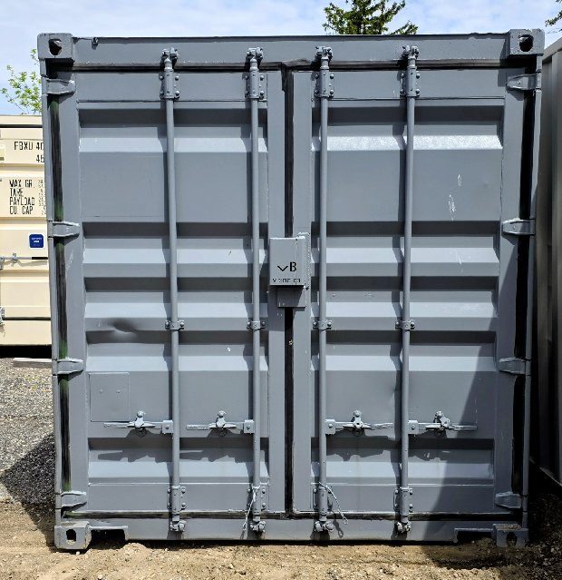 End-on view of a repainted grey 12 foot container with four locking bars, a VB-marked lockbox on the centre seam and corner castings, parked on gravel under a blue sky