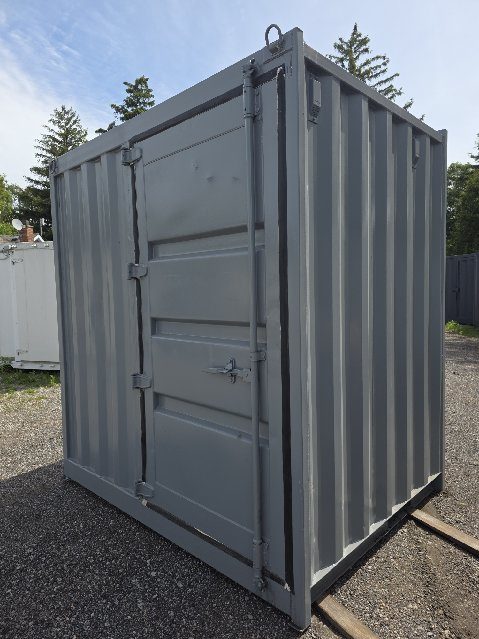 Grey 5 foot 10 inch steel box with one oversized hinged door on its end, resting on timbers in a gravel lot