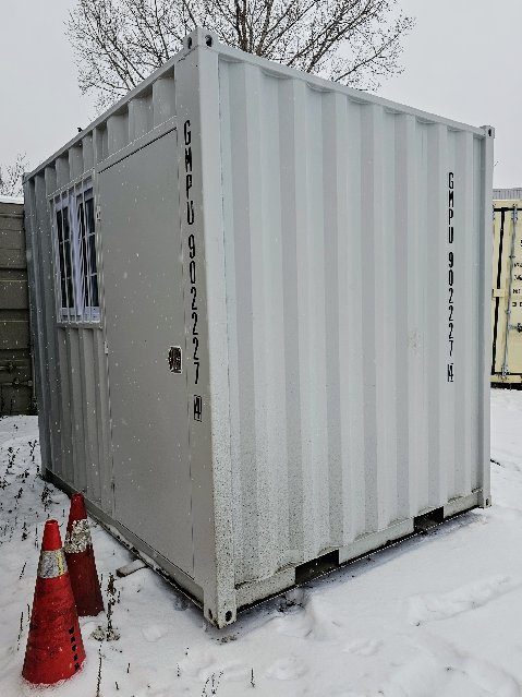 Blank end wall of a grey 9ft standing in snow, with two fork pockets in its bottom rail, a man door and barred window on the side and orange cones by the corner