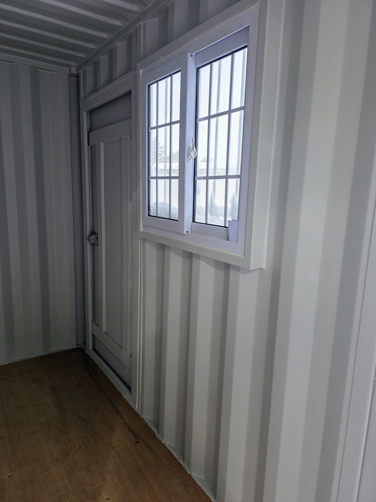 Inside a light grey 9ft container: a white sliding window with bars outside, a closed man door beside it and a plywood floor under white corrugated walls
