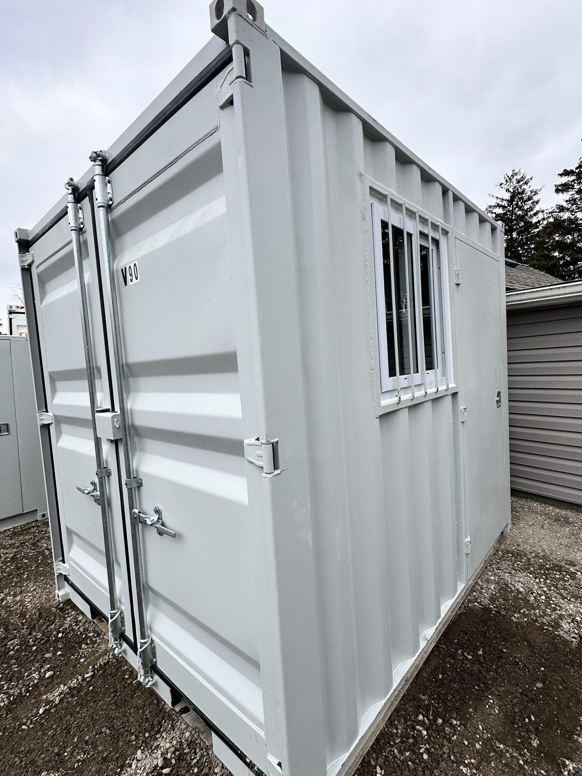 Grey 9 foot container on packed dirt with galvanized locking bars on its cargo doors and a barred sliding window beside a man door on the side wall