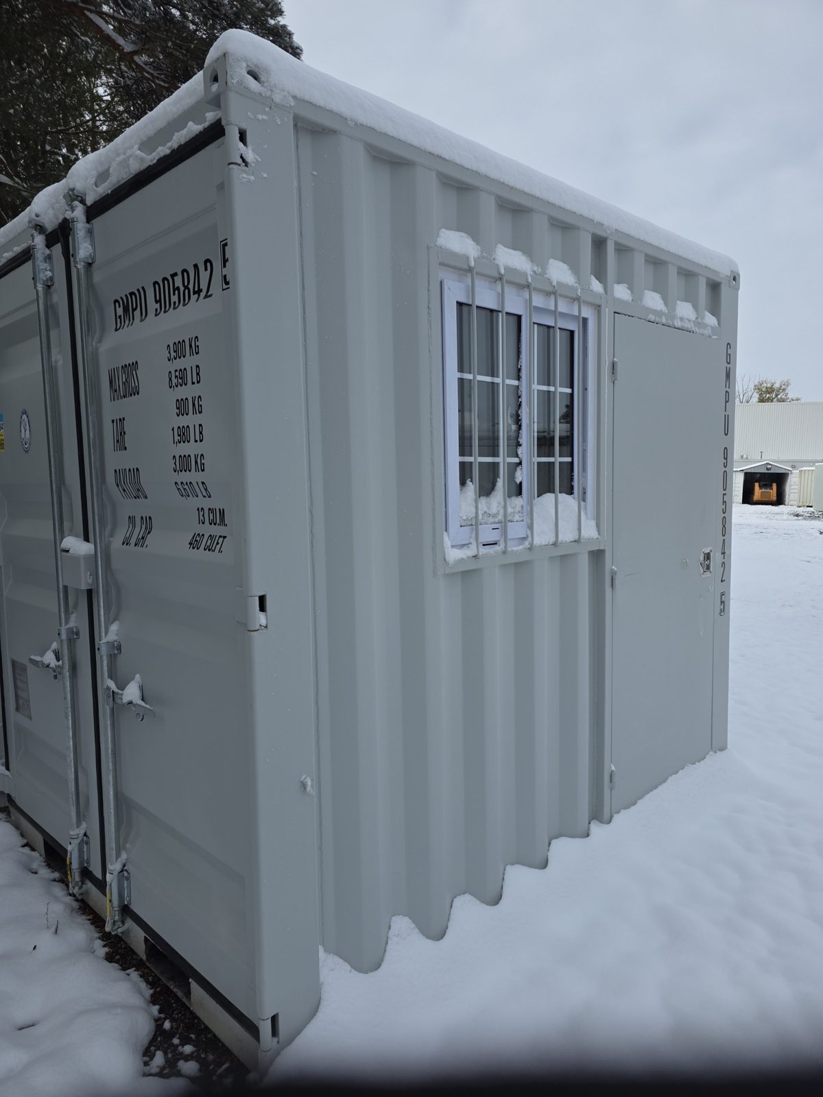 Snow on a light grey 9ft container with weight ratings painted on its cargo door, a barred window and a man door in the side wall