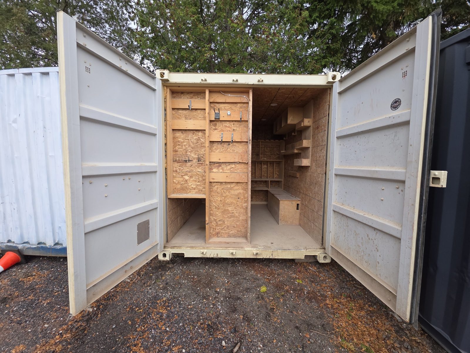 Beige container with both cargo doors swung open on a gravel lot, a framed OSB partition with hooks just inside and shelving further back
