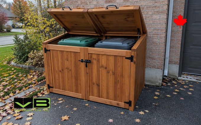 A cedar garbage bin enclosure with both lift-up lids open, showing a green organics cart and a grey garbage cart inside, beside a driveway scattered with fallen maple leaves and a brick wall behind