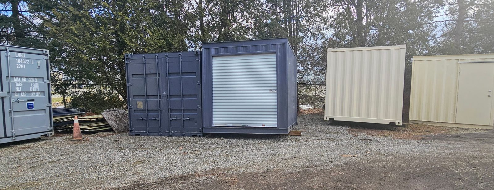 Charcoal 10ft with a white roll-up door parked next to a charcoal container with cargo doors on a gravel lot at a Van Blanc yard, beige units and spruce trees behind