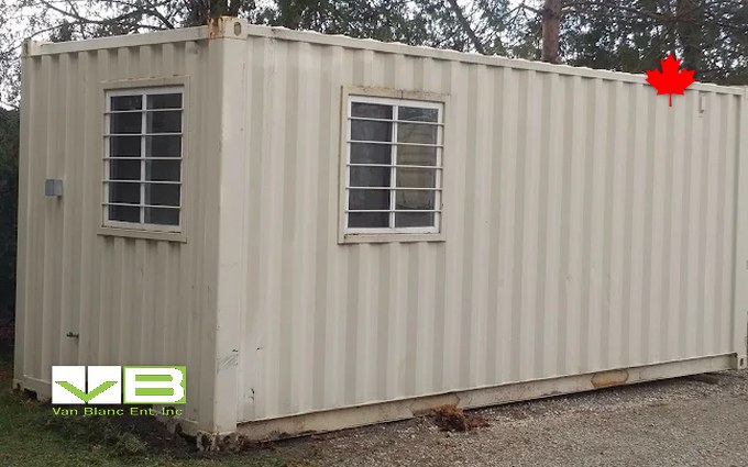 A tan 20-foot container site office with two barred windows and a wall-mounted light, set on gravel among evergreens