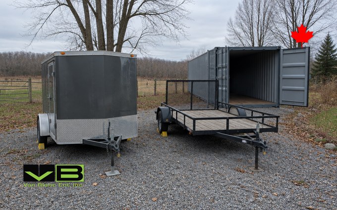 A small enclosed cargo trailer and an open single-axle utility trailer parked on gravel in front of a grey shipping container with its doors open, with bare trees and a farm fence behind