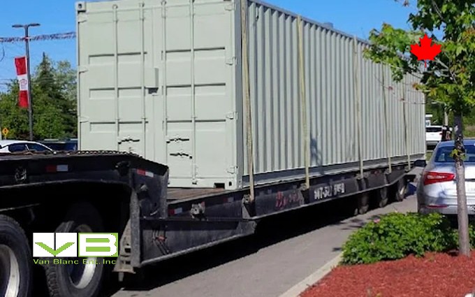 A pale green unit strapped to a flatbed trailer, arriving along a tree-lined residential street.