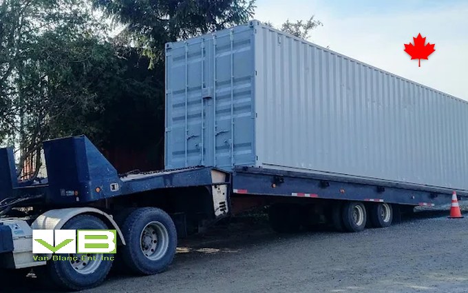 A grey 40-foot shipping container riding on a tilt-deck delivery trailer, parked on gravel beside a tree with a pylon by the rear wheels