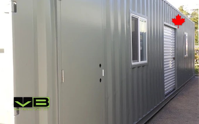 A grey 40-foot shipping container fitted with a steel personnel door and a window on its long side, the lockable feed and medicine room beside a livestock shelter