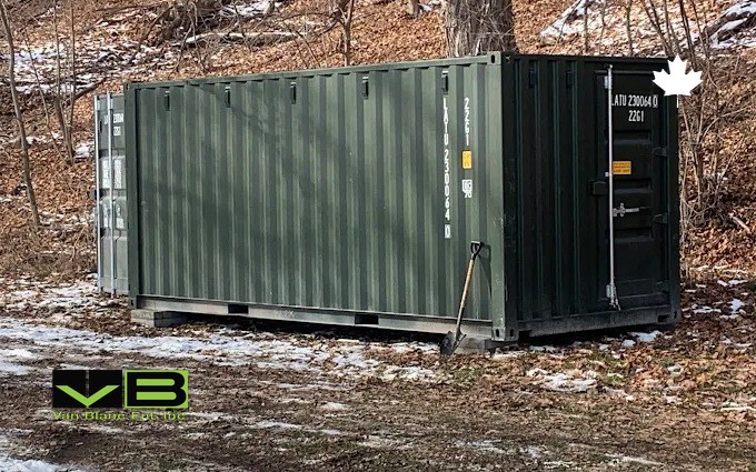 A green 20-foot container placed at the edge of a wooded lot with the first snow of the season on the ground