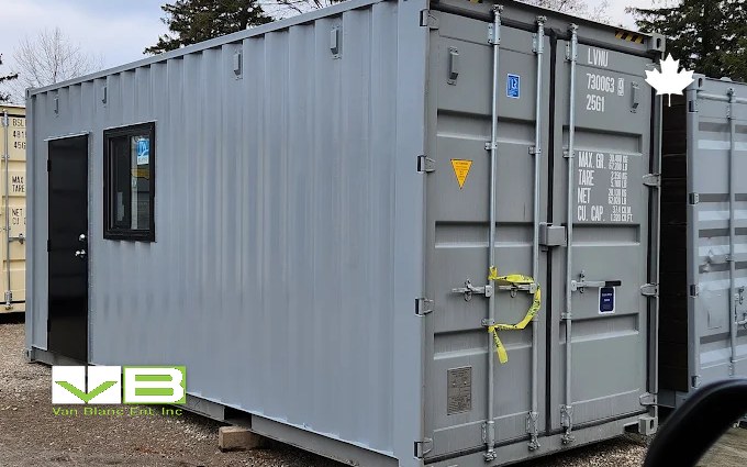 A grey 20-foot shipping container fitted with a black personnel door and a small window, standing on gravel beside other containers at the yard