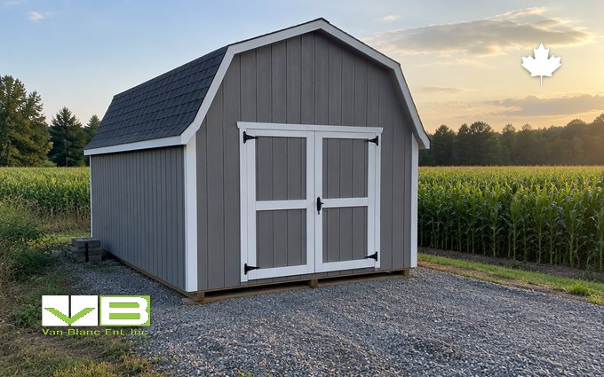 A grey board-and-batten storage shed with a gambrel roof, white trim and double doors on a gravel pad beside a cornfield at sunset