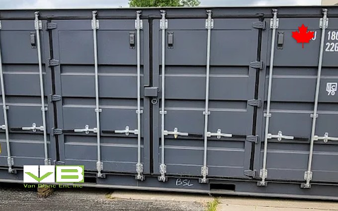 A grey 20-foot open-side shipping container with bi-fold doors running the full length of one long wall, parked on asphalt beside a fence