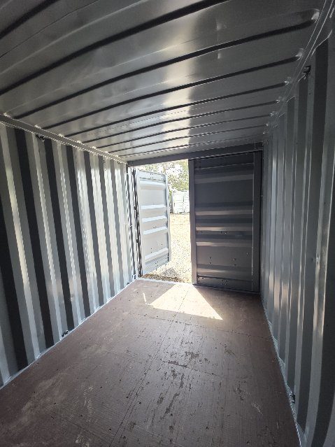 View from the back wall of the 12ft toward its doors, one door open to a gravel yard and other containers, the grey painted walls lit by the sun