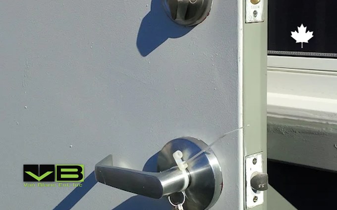 Keys hanging from the lever handle of a shipping container's grey steel personnel door, with a deadbolt above and the strike plates showing on the open frame