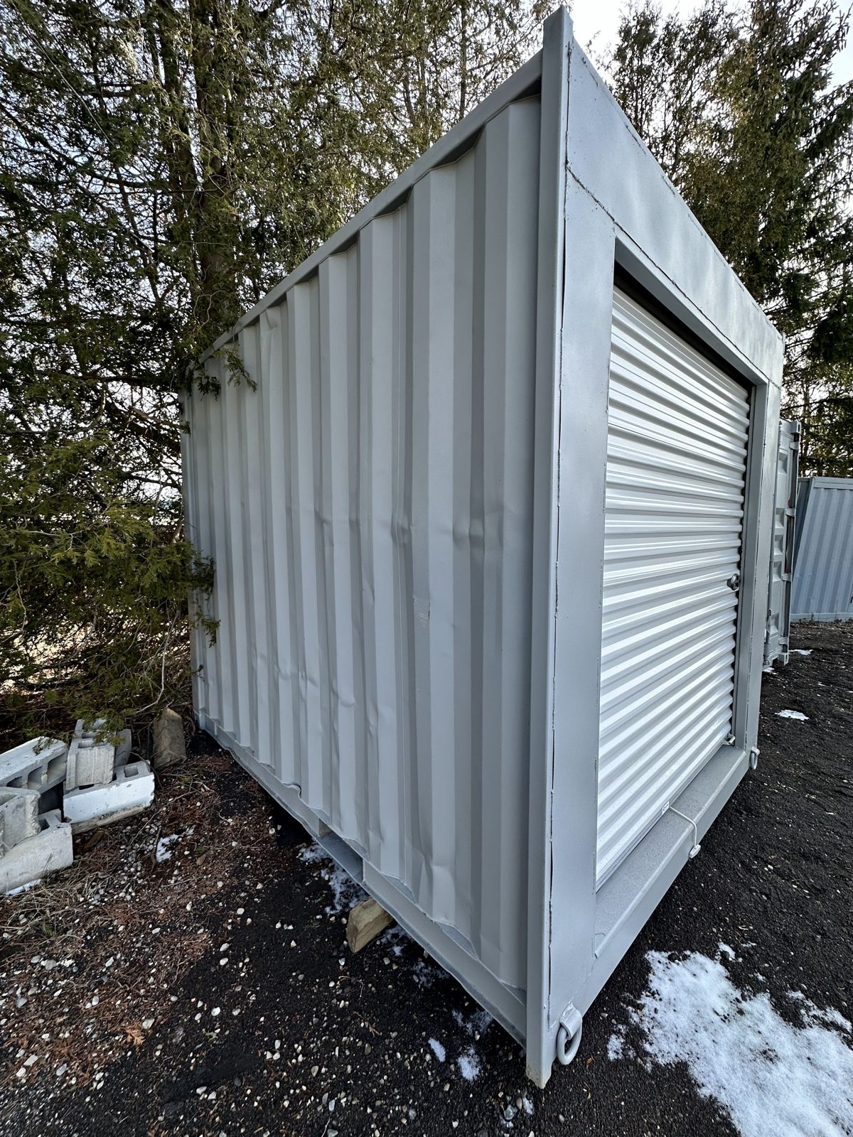 Side view of a repainted light grey 10ft container, corrugated wall along the left and a white roll-up door filling the end, resting on a wood block beside spruce trees