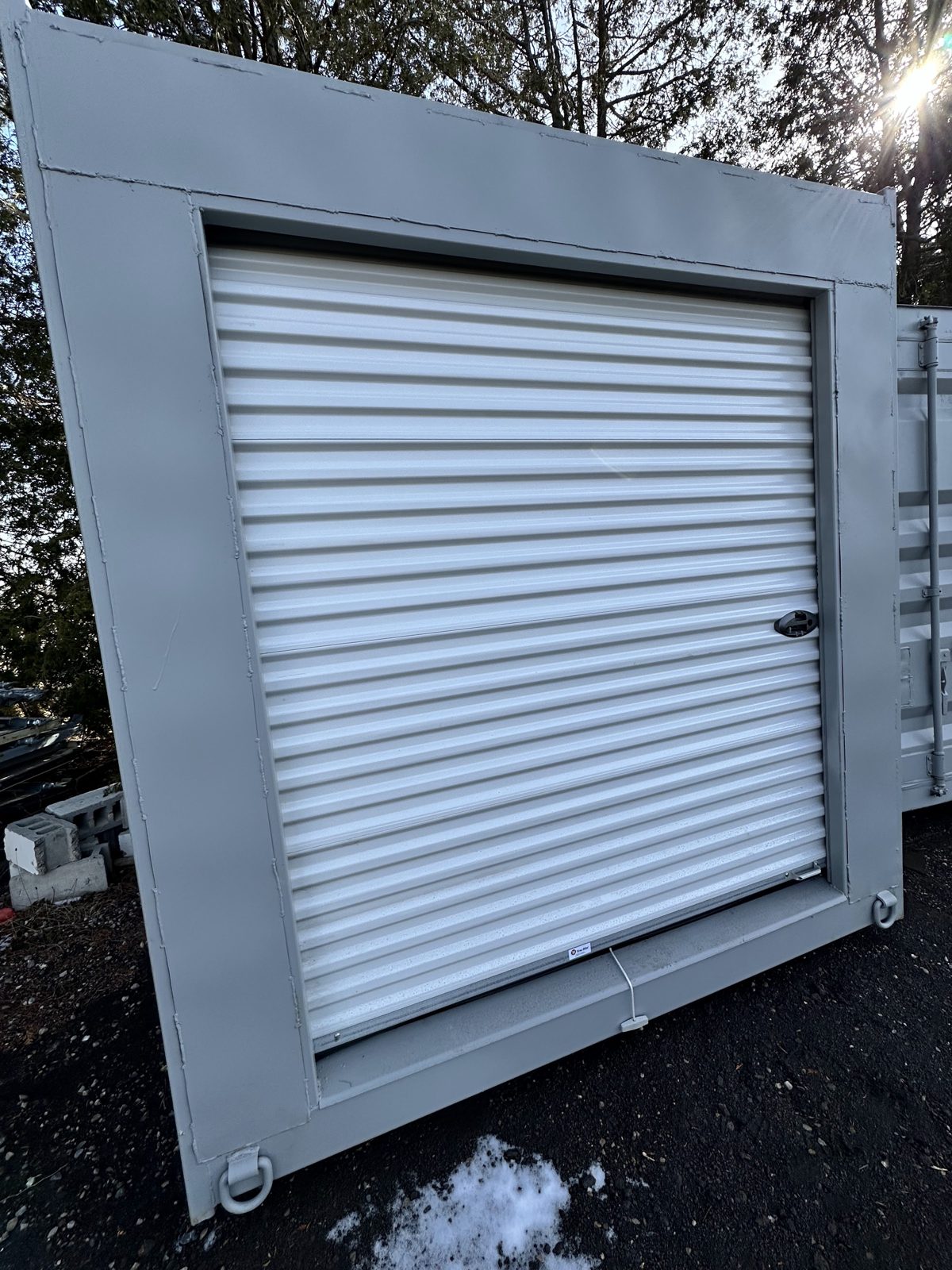 Closed white roll-up door with a side latch, set in a flat steel frame across the end of a light grey container, with a patch of snow on the dark ground in front
