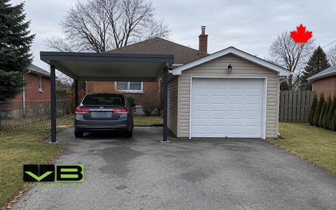 An open two-post carport with a flat roof sheltering a grey sedan beside a small single-car garage with beige vinyl siding and a white overhead door, on a paved driveway in front of a brick bungalow
