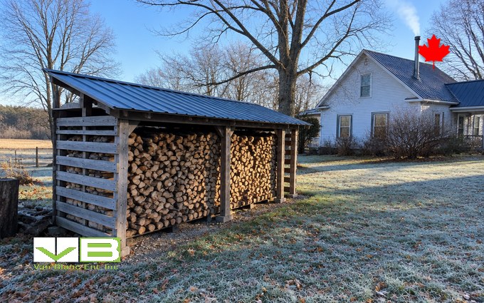 An open-fronted firewood shed with a sloped steel roof and slatted sides, stacked full of split hardwood, beside a white farmhouse with frost on the lawn and bare maples behind
