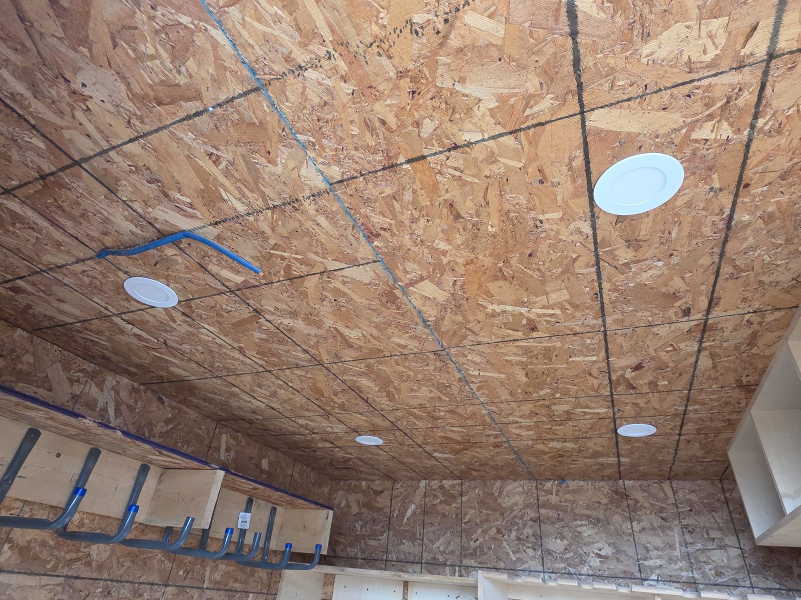 Looking up at an OSB ceiling with four round flush lights, a rail of blue-tipped steel hooks along the upper wall and a shelf unit at the right edge