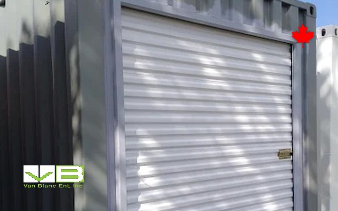 A white roll-up door framed into the grey corrugated wall of a converted unit, dappled in tree shade.