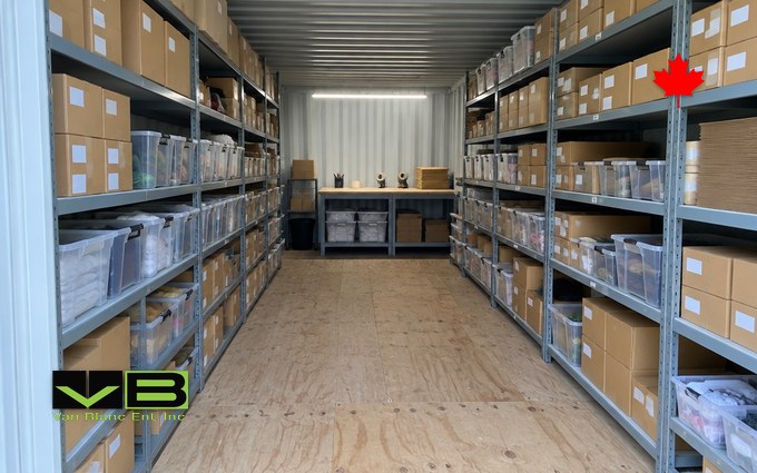 Looking down the inside of a shipping container fitted as a stock room, with steel shelving of labelled boxes and clear bins along both walls and a packing bench at the far end under a strip light