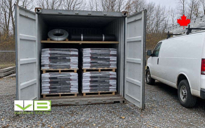 A grey 20-foot shipping container with its doors open on a gravel yard, wrapped bundles of asphalt shingles stacked on wooden pallets inside with a coil of drip edge and a roll of underlayment on a shelf above, and a white work van with a ladder on the roof rack parked beside it