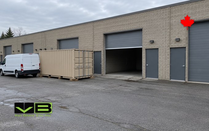 A small multi-tenant industrial plaza bay with a grey drive-in door and a man door, a for-lease sign removed, and a beige 20-foot shipping container on blocks in the shared yard beside a parked cargo van
