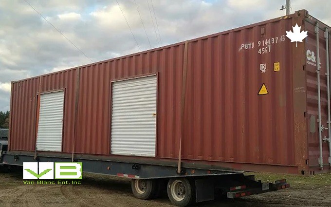A rust-red forty-foot container with two white roll-up doors riding a flatbed trailer into a rural yard