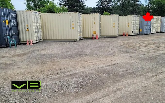 A long row of tan and grey 20-foot storage containers on a gravel lot at the Brantford yard, orange pylons between them and trees behind