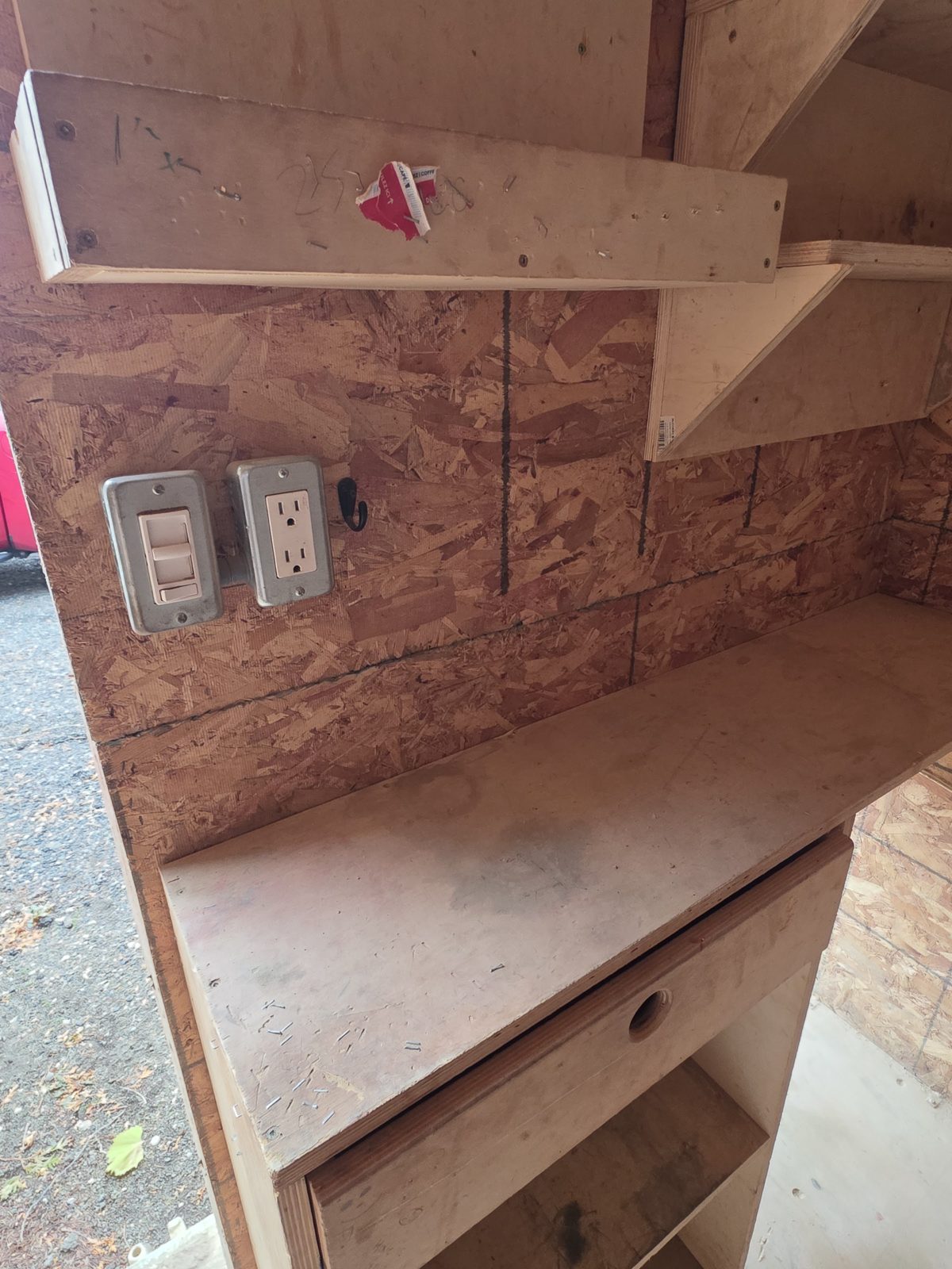 Wall switch and duplex outlet in metal cover plates above a plywood workbench with a drawer, beside the open cargo door