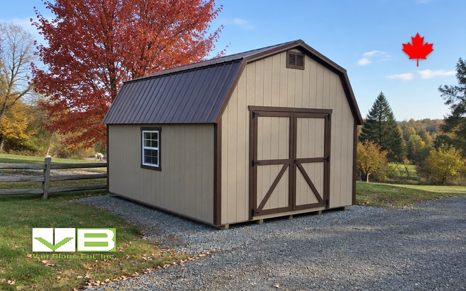 A tan lofted barn-style shed with a brown metal gambrel roof, brown trim and double doors set on a gravel pad beside a split-rail fence, with a red maple and a field behind