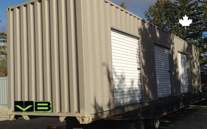 A tan shipping container fitted with white roll-up doors arriving on a flatbed trailer, the steel replacement for a storage tent