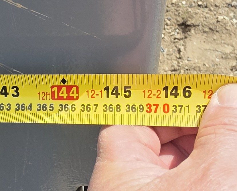 Hand holding a yellow tape measure against the end of grey container steel, the steel edge near the 146 inch mark and the 370 cm tick, with the 12ft label at 144 inches to the left