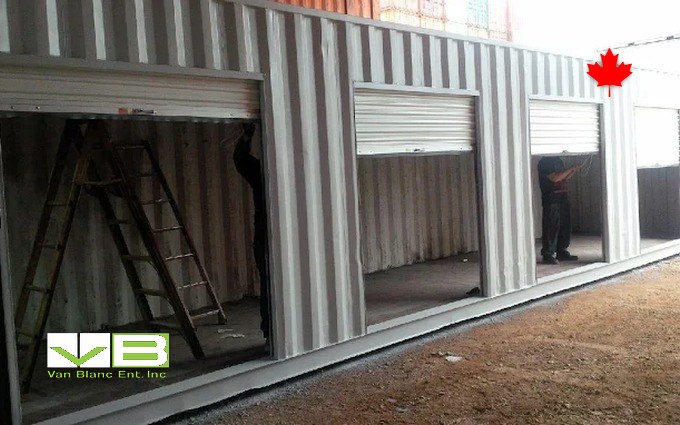Roll-up door bays being fitted into a row of containers, ladders and crew mid-install