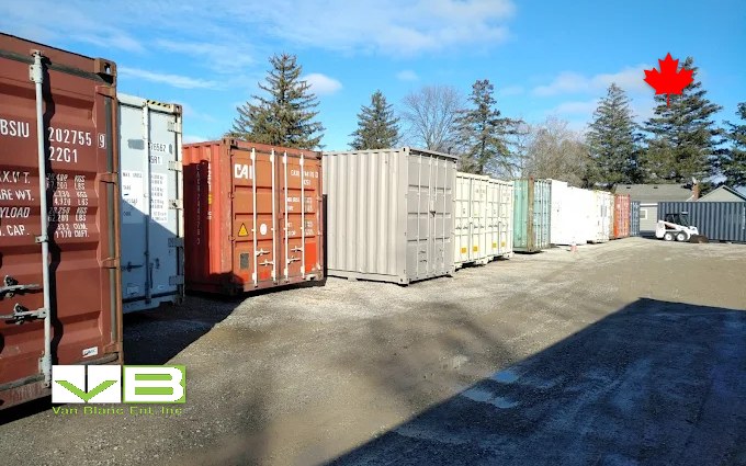 A row of used shipping containers in red, white, tan and green standing on gravel at the Brantford yard on a bright winter day, a skid steer parked at the far end