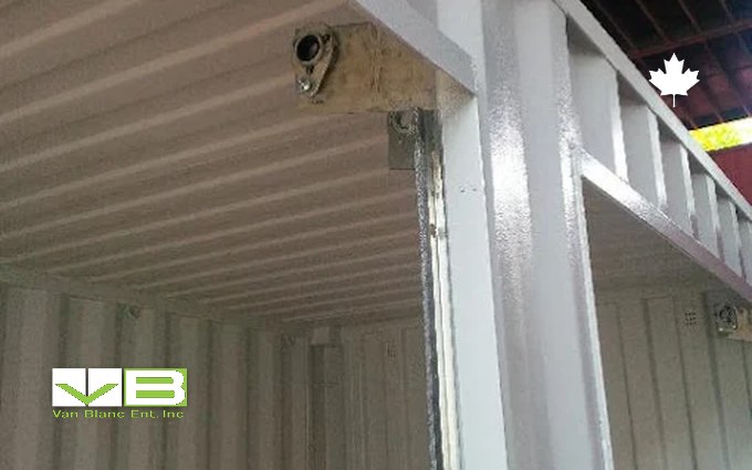 Looking up inside a white-painted shipping container conversion at a round vent mounted in the ceiling corner beside a steel post, with the open doorway beyond
