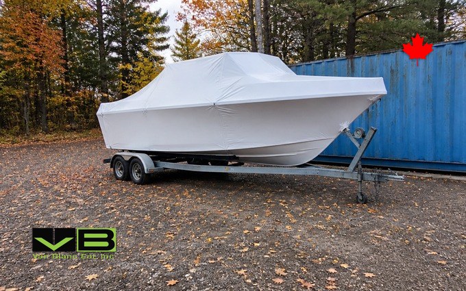 A shrink-wrapped boat on its trailer parked on gravel beside a blue container with fall colour behind