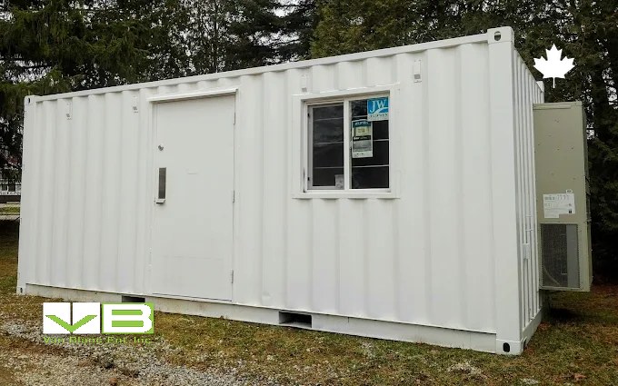 A white 20-foot container office with a personnel door, a sliding window and a wall-mounted air conditioner, set on wood blocks on grass among evergreens