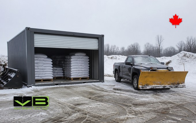 A dark grey steel container with its white roll-up door raised, holding pallets stacked with white bags of road salt, beside a pickup truck fitted with a yellow plow blade in a slushy contractor yard with snowbanks behind