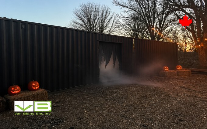 Black painted steel containers set end to end with a ragged cut-out doorway leaking fog, carved pumpkins glowing on hay bales and string lights in the bare trees at dusk