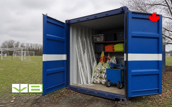A royal blue steel container with a white stripe and its doors open at the edge of a soccer field, holding rolled goal nets, bags of balls, folded pinnies, bins on shelves and a line marking cart, with goalposts and bare trees behind