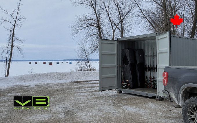 A grey steel container with its doors open on a snowy gravel lot by the shore, holding folded portable ice shelters and rods on racks, a pickup truck beside it and small ice huts scattered across the frozen lake beyond the trees