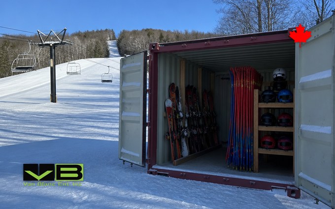 A maroon steel container with its doors open on packed snow at the base of a small ski hill, holding racks of skis and snowboards, bundles of red and blue race gate poles and shelves of helmets, with a chairlift on the slope behind