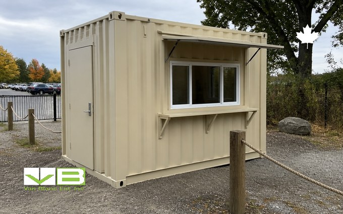 A small cream steel container fitted with a man door, a sliding service window, a metal awning and a counter shelf, at a gravel entrance with rope posts and parked cars beyond in autumn